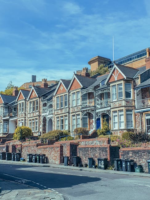 A row of Victorian-style terraced houses on Avenue Road, featuring decorative ironwork balconies, large bay windows, and brick facades, set against a bright blue sky with some scattered clouds. The houses are elevated from the street level, with front gardens bordered by low brick walls and small bushes. At street level, black wheelie bins are lined up along the pavement, and a parked van, partially visible on the right, appears to be used for house removals. The scene depicts an urban residential setting in SWISS COTTAGE, with attention to the architectural details typical of local homes. This image supports topics related to house removals, home relocation, and furniture transport, highlighting the environment where professional moving services like Swiss Cottage removals may operate.